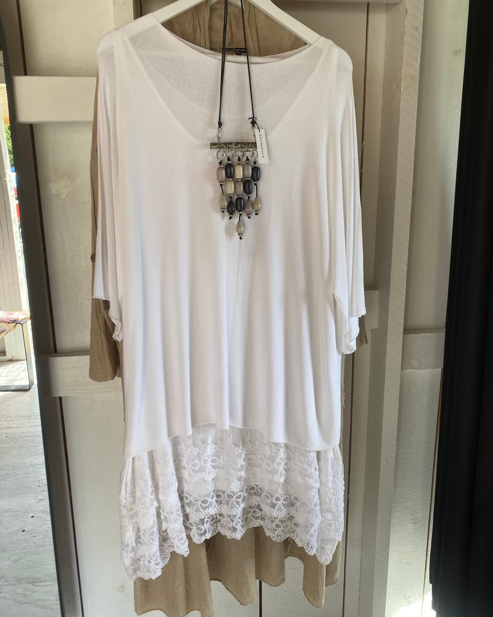 A white lace-hem linen top layered over a tan linen slip dress, with a beaded necklace, hanging against the shed's white plank wall