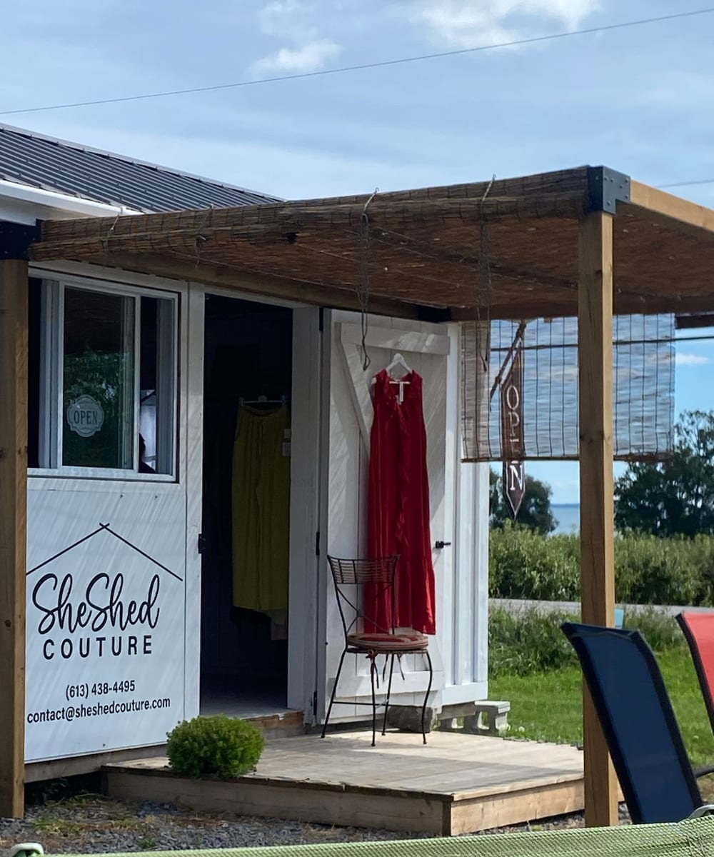 The SheShed Couture shed, with a red dress and yellow linen trousers hanging beside the open door under a reed pergola