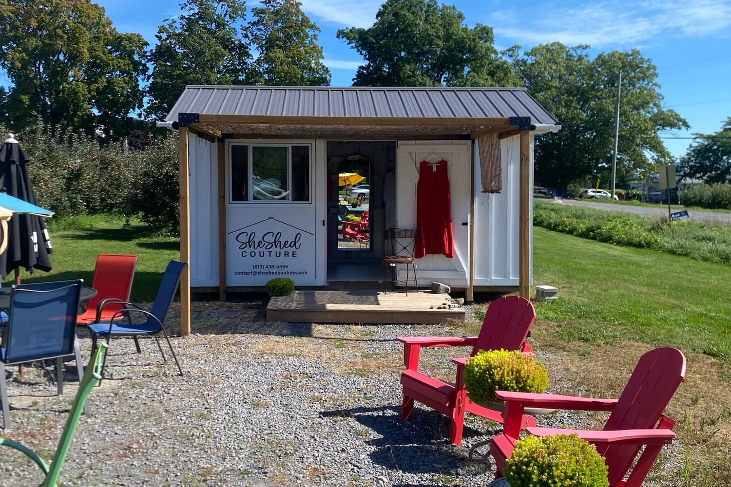 The white shed seen from the gravel lot, with an apple orchard behind it and red Adirondack chairs out front under a blue sky